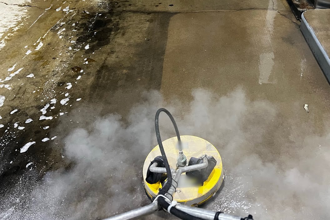 Rotary surface cleaner pressure washing stained concrete beside a fuel-island curb, with mist rising and a clean strip next to the darker, oily concrete