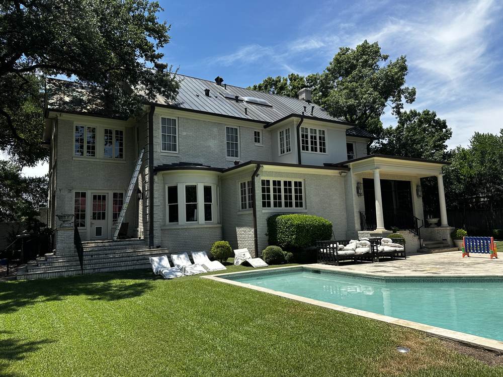 Two-story painted-brick Austin home with a pool and live oaks, ladder set against the wall during exterior cleaning