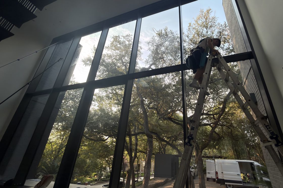 Casa Care technician on a ladder cleaning the inside of a tall wall of black-framed windows on a commercial post-construction job, with live oaks, a brick wall and a parked white van outside